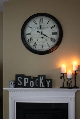 A clock on the wall above a fireplace mantel with the words Spooky on the mantel.