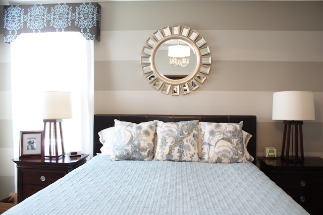 A baby blue bedspread, throw pillows on the bed, a striped wall and a round mirror.