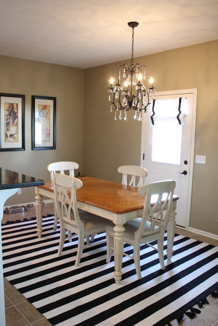 A striped rug, wooden table with white chairs and the chandelier hanging up in the dining room.
