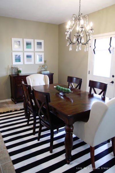 Dining Room with black and white striped rug, wood table, linen chairs, iron and crystal chandelier and beach photograph art.