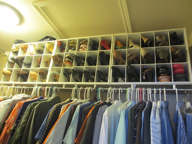 Men's shirts hanging in the closet with shoes in cubby holes above the shirts.