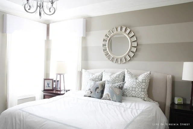 Bedroom with striped walls, white bedding, blue and gray pillows and chandelier.