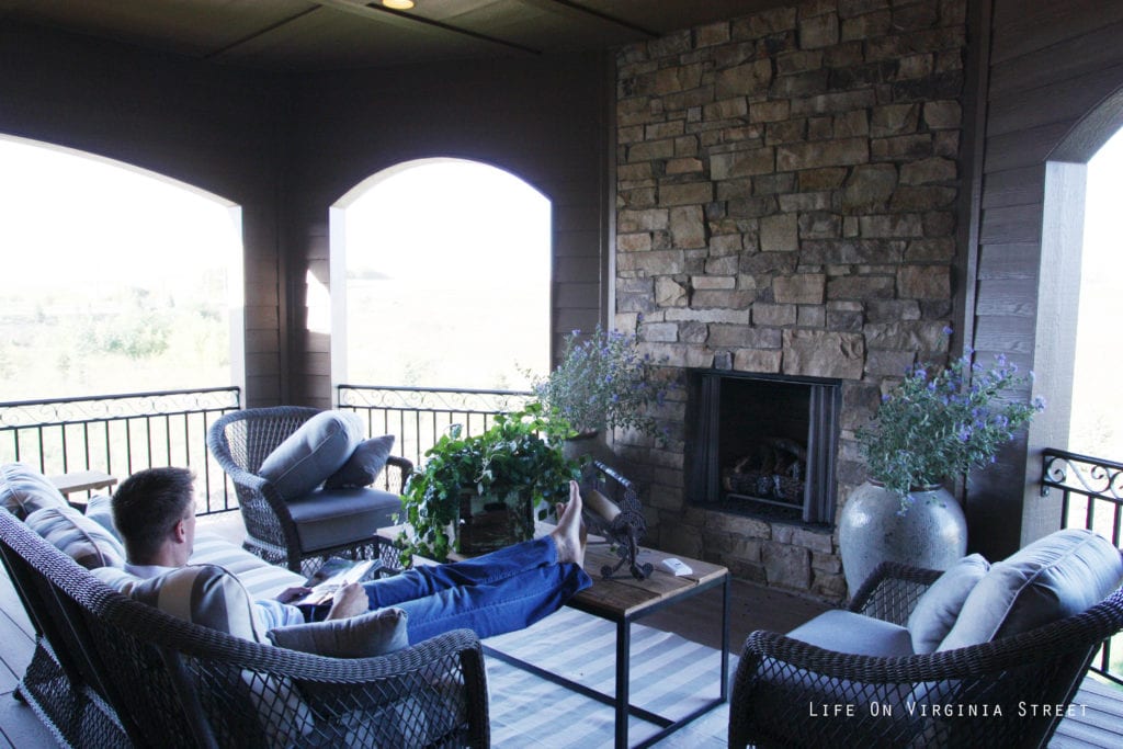 A man sitting in the outdoor patio with a fireplace and comfy chairs.