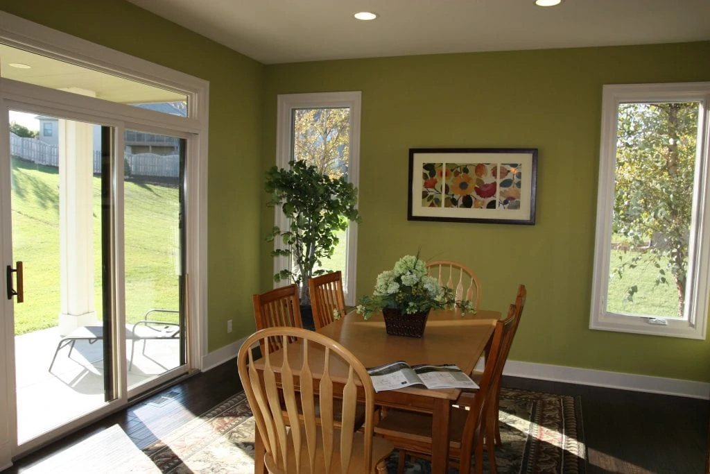 A sunny dining room with a light colored wood table, light green walls, and sliding door leading onto a patio.