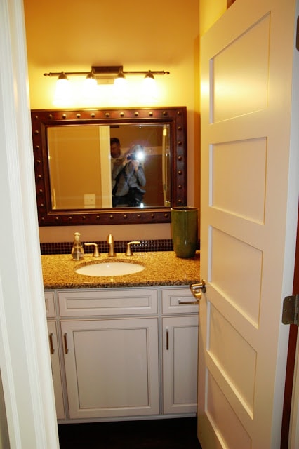 The bathroom with an open door and large mirror.  White cabinets on the bottom.