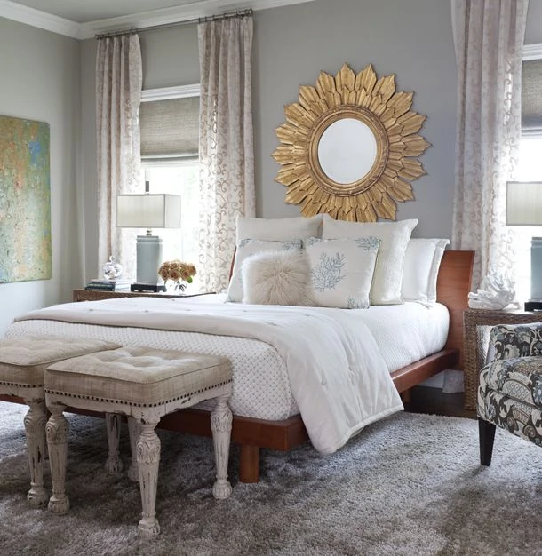 Large sunburst gold mirror on blue/gray wall with bed underneath and white bedding. Tufted footstools are in front of the bed.