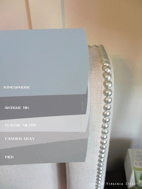 Side-by-side comparison of Behr Atmospheric, Behr Pier, Behr Fashion Gray, Behr Classic Silver, Behr Antique Tin next to an upholstered chair which makes the colors look more toned-down.
