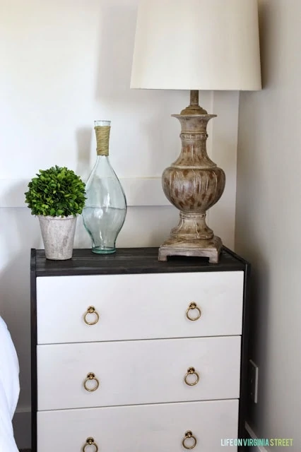 A nightstand with a ceramic lamp and white shade on it.  A green topiary and clear glass vase.