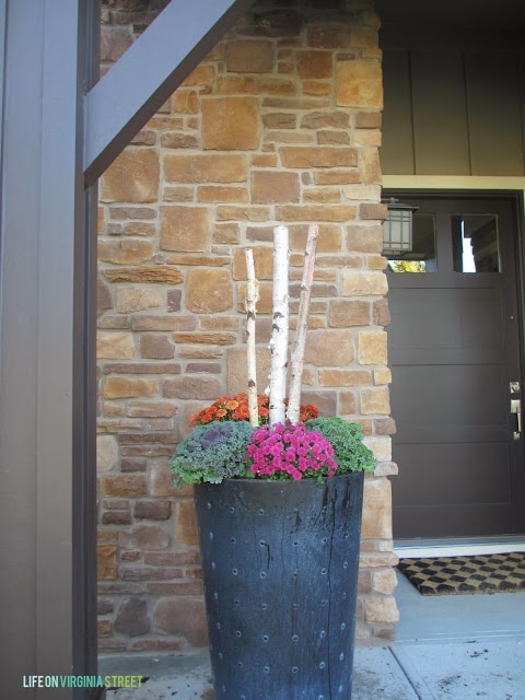 Large planter with kale and purple flowers and branches beside stone house.