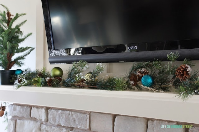 Black tv above the mantel with blue, green Christmas balls and greenery around it