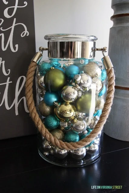 A clear glass jar is filled with Christmas ornaments in blue, green and silver.