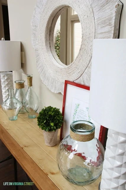 There is a white mirror above a wooden console table filled with clear glass.