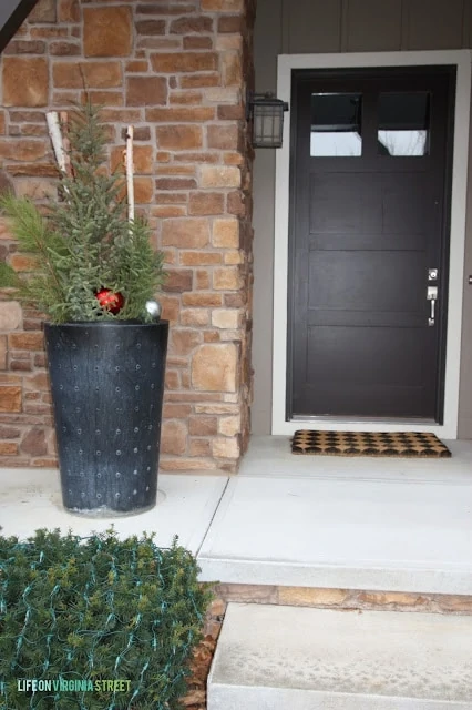 There is a planter on the front porch with a Christmas tree in it decorated with ornaments.