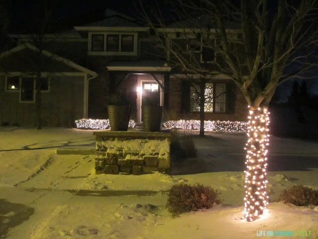 The outside of the house with snow on the ground and trees with lights.