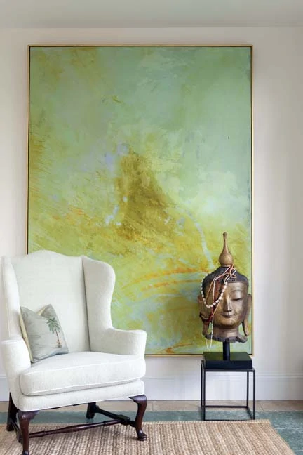  A very large green, yellow and soft blue painting hanging on a wall with a white chair and a Buddha head in front of it.