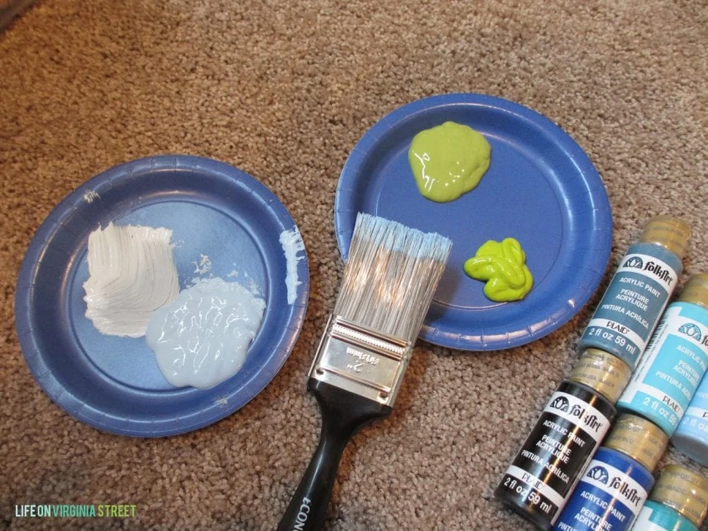 Blue paper plates with white, paint on one and yellow paint on another with a paint brush beside it.