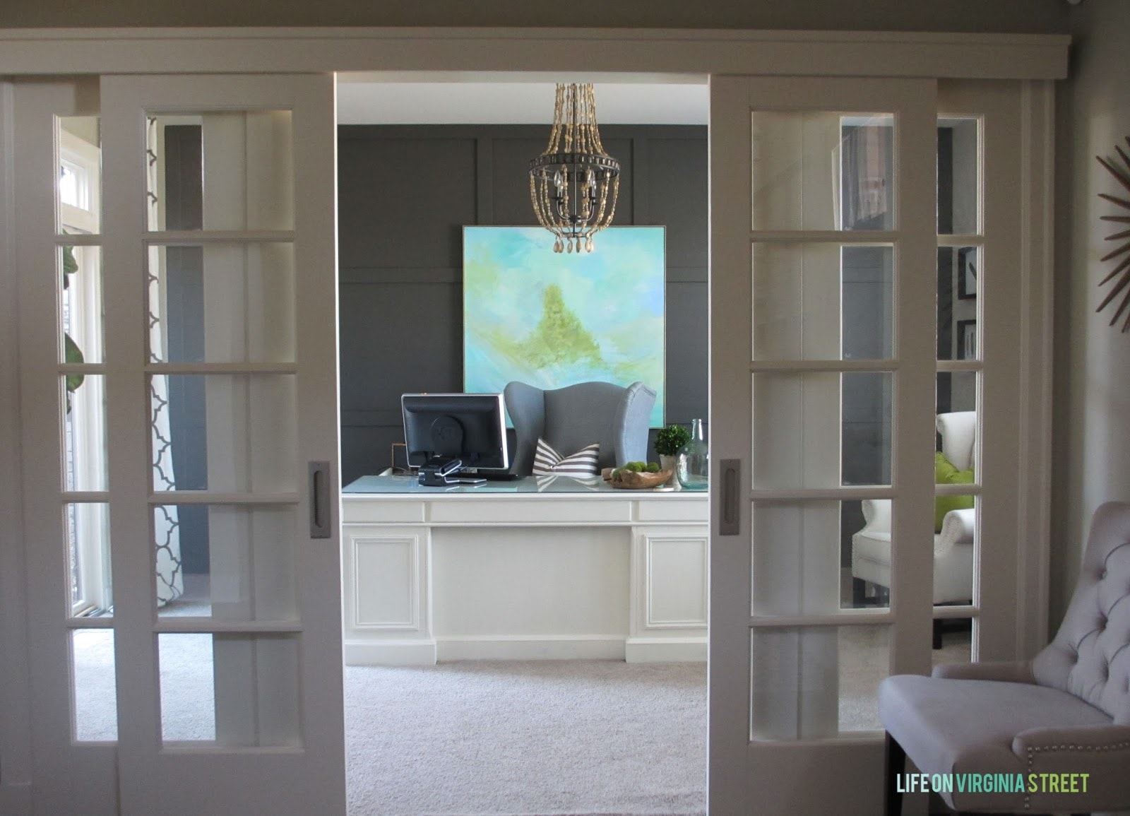 Sliding office doors leading to a home office with Urbane Bronze painted board and batten wall, large abstract art, a white desk, a wood bead chandelier and slate blue office chair.