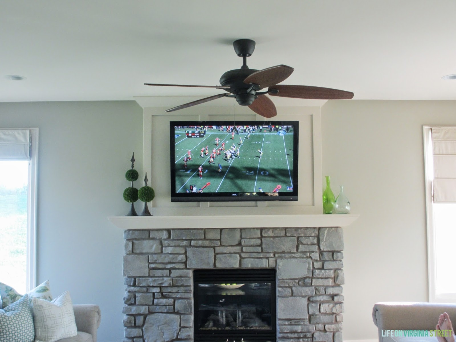The fireplace with the TV above it and a football game on the TV. A ceiling fan hanging in the room.