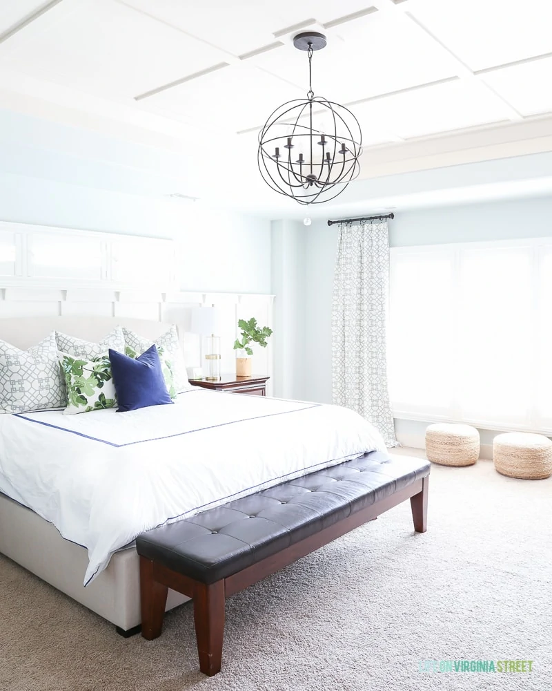 Master bedroom with Sherwin Williams Sea Salt walls, board and batten ceiling, white bedding, navy blue velvet pillow, fig leaf pillows, jute poufs and trellis curtains.