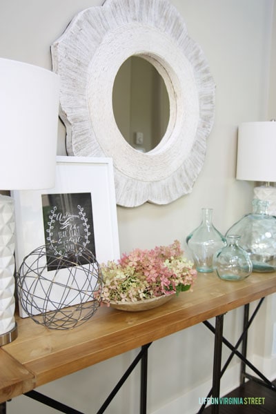 White mirror above a wooden side table with unique glass vases on it.