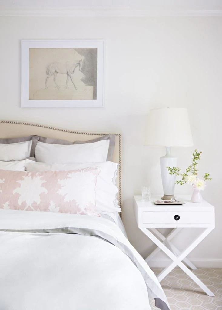 A white bedroom with a pink and white pillow and light pink vase on the nightstand.