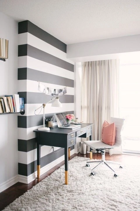 Black and white striped walls in office with a black desk in front of it and a white chair with a pink pillow on it.