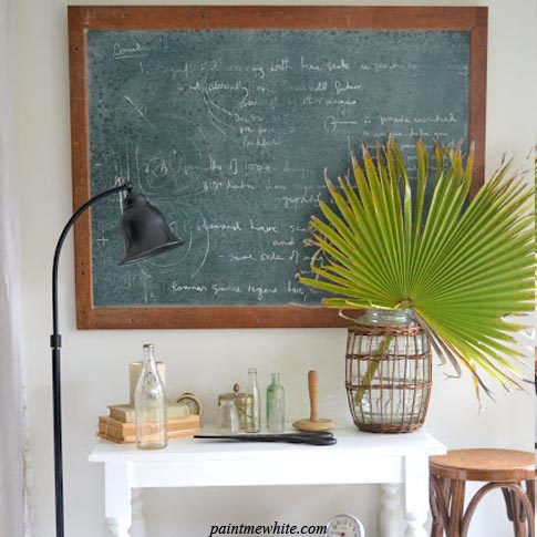 A white side table with a chalkboard above it and a palm frond on the table.