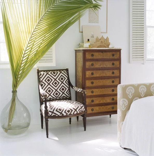 A large palm frond in a clear glass vase on the floor beside a chair and dresser.
