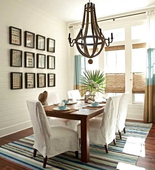 A coastal style dining room with a striped rug, white shiplap walls, wine barrel wood chandelier, palm fronds, gallery wall, woven shades and striped curtains. 
