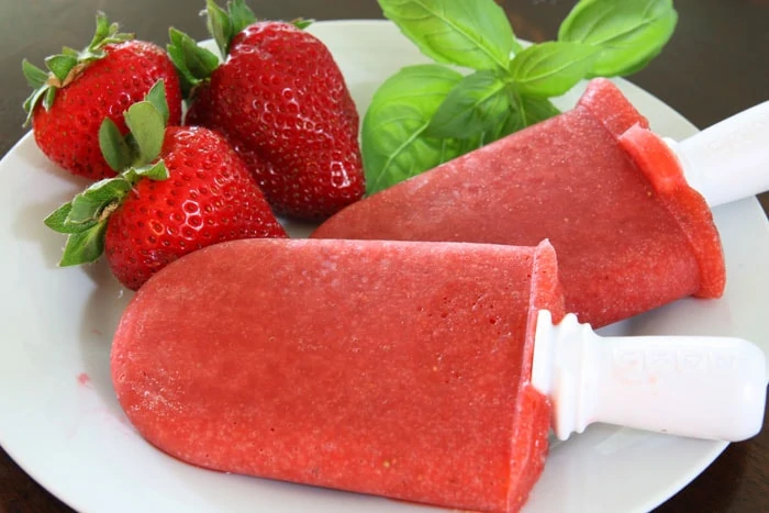 Two Strawberry Basil popsicles on a plate with a couple of fresh strawberries beside them.