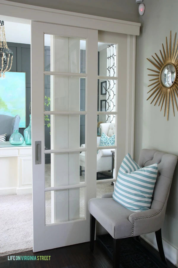 White sliding doors leading into office with grey chair outside and a mirror on wall.