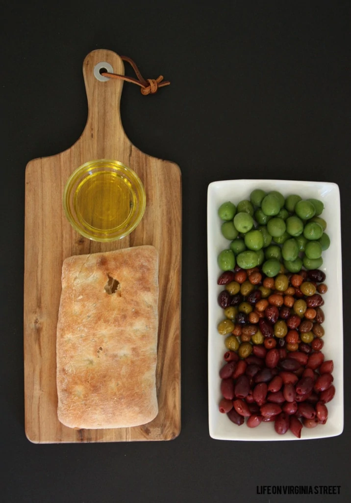 Bread board with bread on it, and olive oil for dipping, plus a tray of olives beside it.