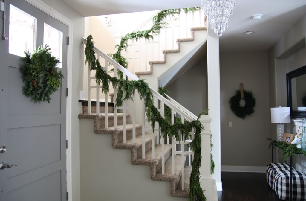 A beautiful Christmas entryway with fresh evergreen garland, a pinecone wreath, black and white buffalo check poufs, and a crystal chandelier.