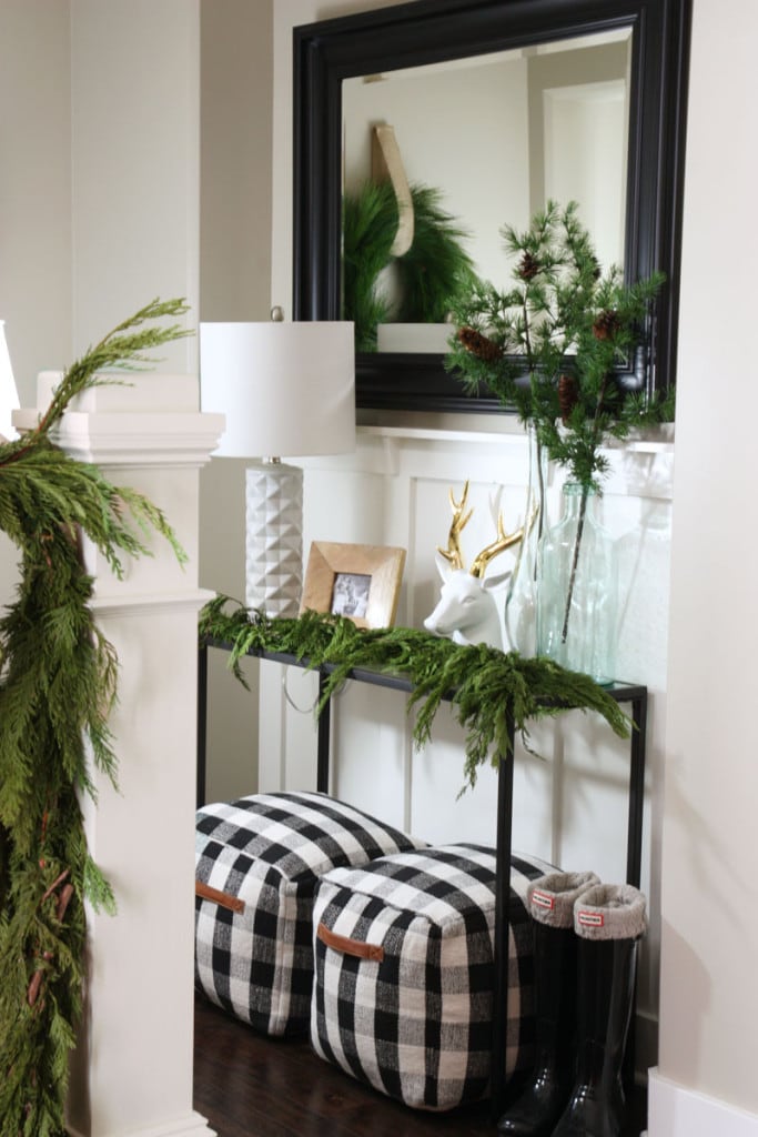 A darling Christmas entryway with a black iron console table, black and white buffalo check pouf ottomans, a black framed mirror, fresh garland swag, and black, white and green accents.