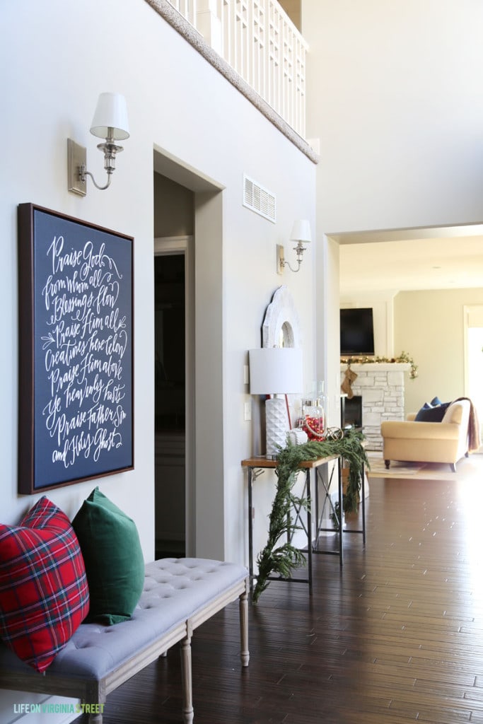 Beautiful Christmas hallway featuring a Doxology canvas hanging above a gray linen bench with a red plaid and green velvet pillow.