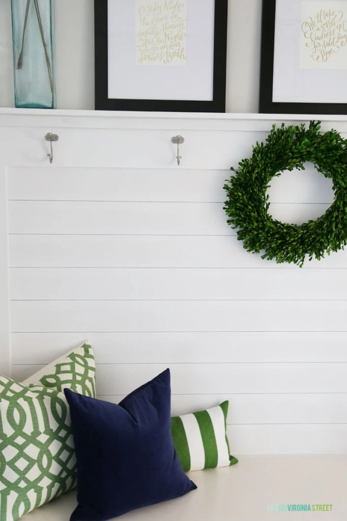 A Christmas mudroom with green trellis pillow, navy blue velvet pillow, a boxwood wreath, and Christmas hymn artwork.