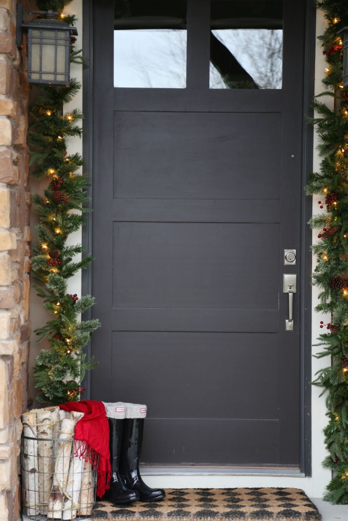 Front Door Christmas Decor including garland swag, birch logs in a wire basket, a red scarf, and black hunter boots with cableknit boot socks.