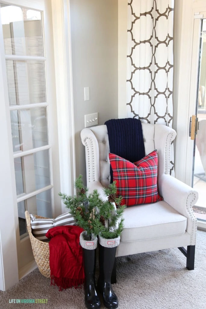 Office Christmas Chair with red plaid pillow, black glossy Hunter boots filled with greenery, and a trellis curtain panel.