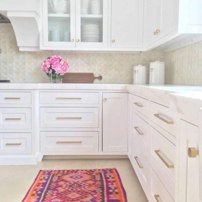 An all white kitchen with pops of pink flowers and a pink rug with orange and purple in it.