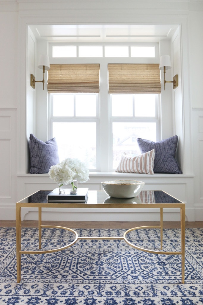 Gold table with glass top. White flowers on top and blue pillows by the window.