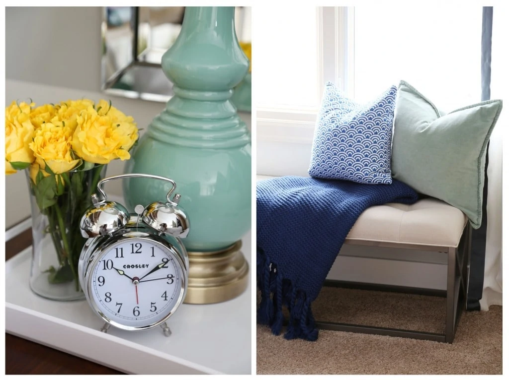 Am alarm clock on the nightstand, yellow flowers and a light green lamp. There is a settee by the window with a blue blanket and pillows.
