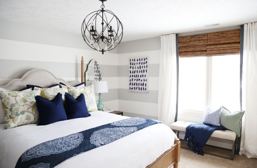 A round metal chandelier above the bed with blue pillows and blanket on the bed and a window seat.