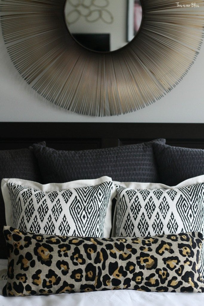 A master bedroom with a gray headboard, gray, black pillows and a large metallic mirror above the bed.