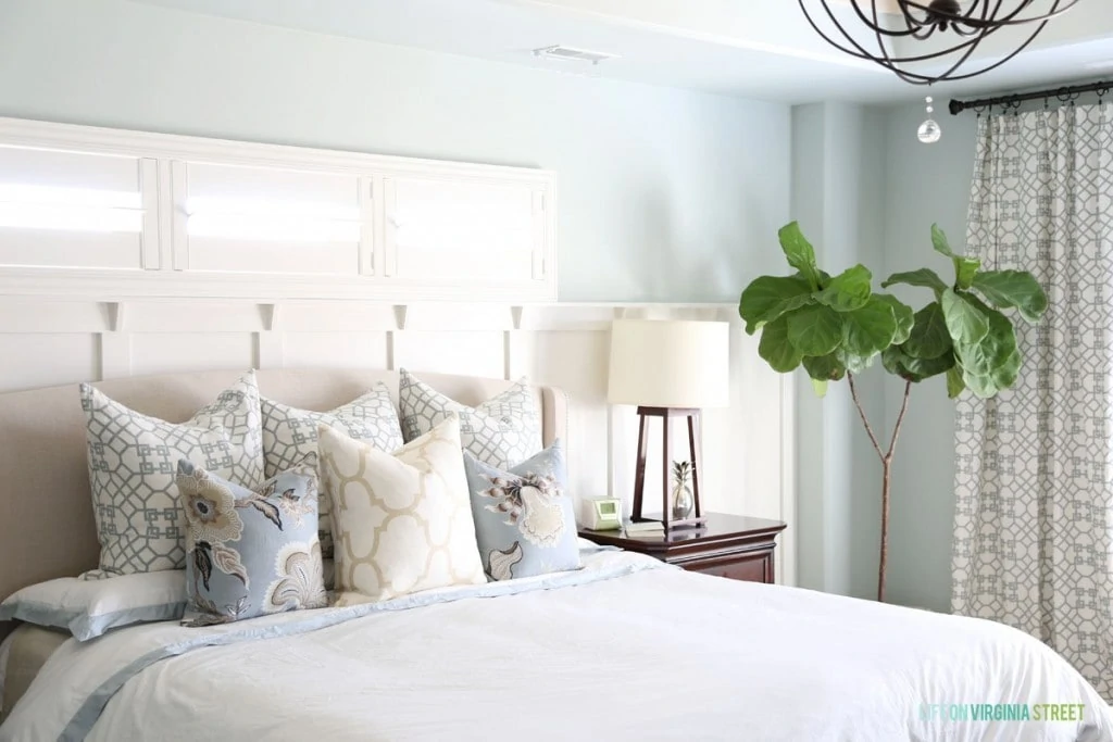 A master bedroom with a large bed, pillows on the bed, a wooden side table with a lamp, and a green plant in the corner of the room.