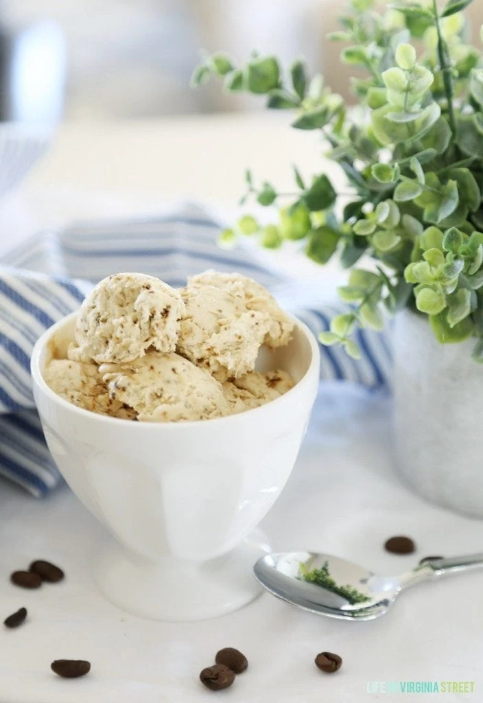 Coffee ice cream in a white bowl with a spoon beside it and coffee beans on the counter beside it.