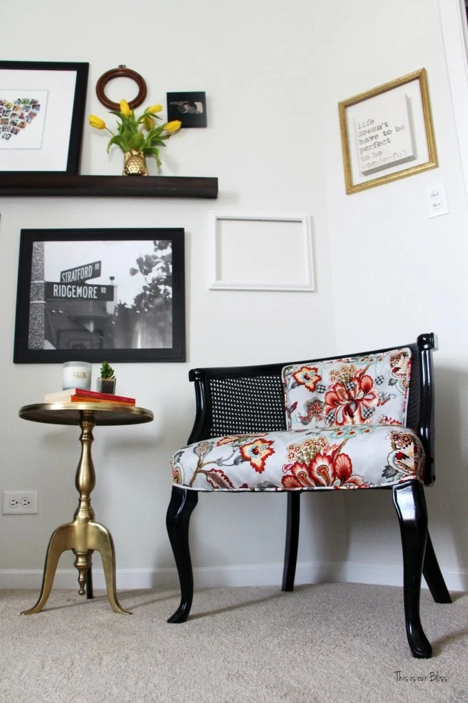 The corner of a room with a black chair covered a floral design on the seat cushion, and a gold table and on a wall shelf a pineapple vase filled with yellow tulips.