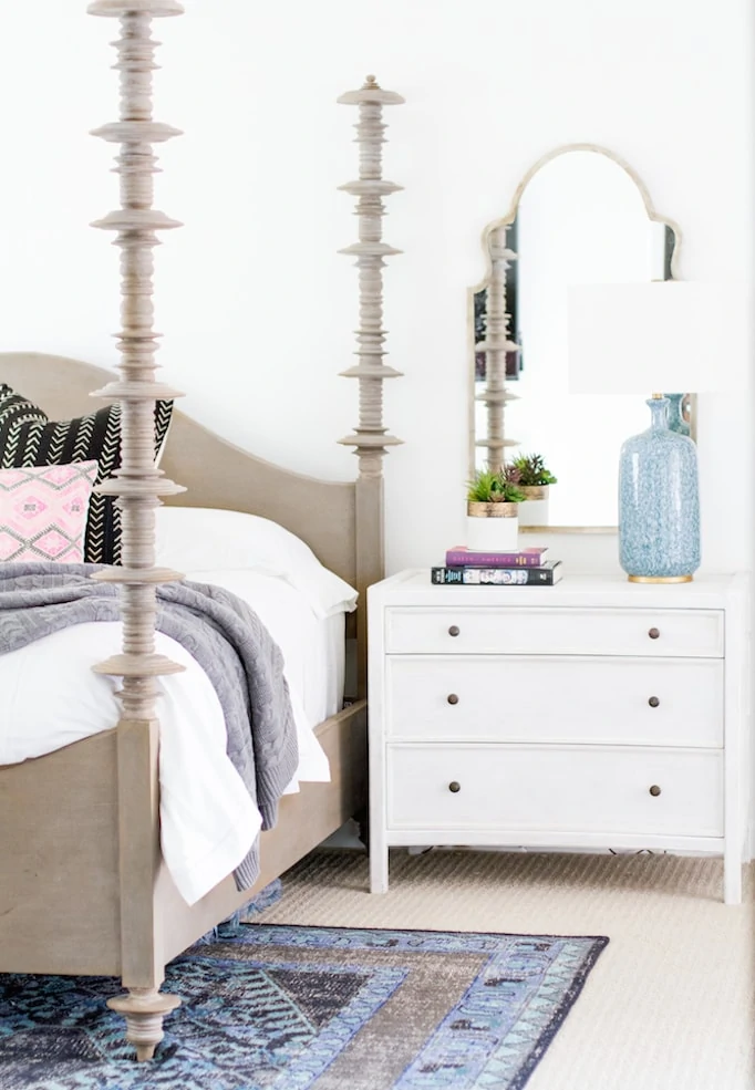 In a medium sized bedroom there is a patterned blue rug, a white side table with drawers and a mirror on the wall.