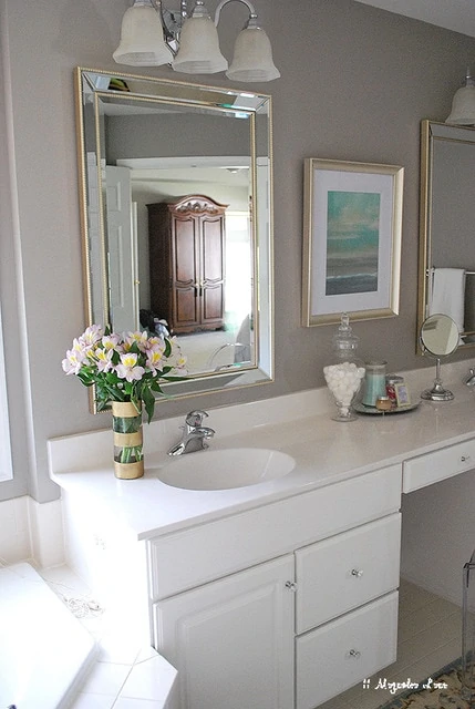 A bathroom with white cabinets,mirrors rimmed with gold and flowers on the white counter.