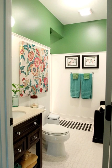 A small bathroom with an wall half green, a floral print on the wall and a dark wood cabinet with a white counter.