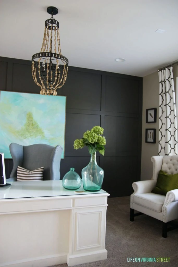 A coastal home office painted Sherwin Williams Urbane bronze, with a white desek, colorful abstract art, trellis curtains, and a wood bead chandelier.